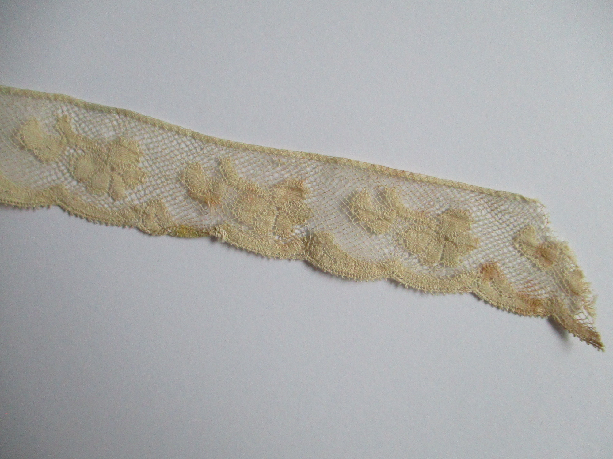 Length of folded silk lace trim.: End detail of folded cream silk lace trim. (i)